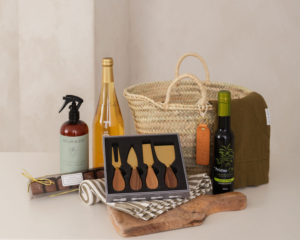 Gourmet gift basket with wine, cheese tools, and other items on a neutral background